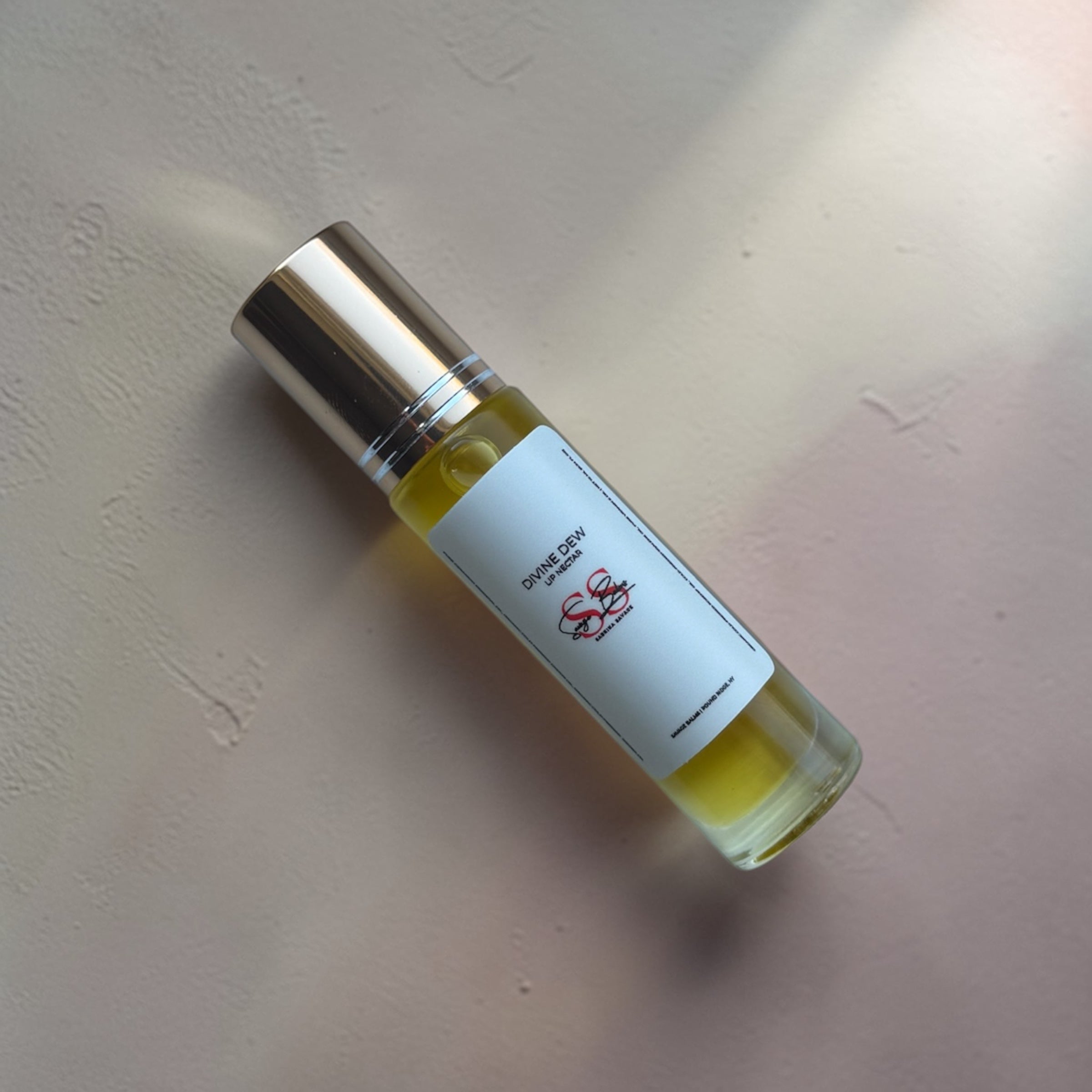 Small lip oil bottle with a gold cap and white label on a beige surface, labelled "Divine Dew Lip Oil" by Savage Balms