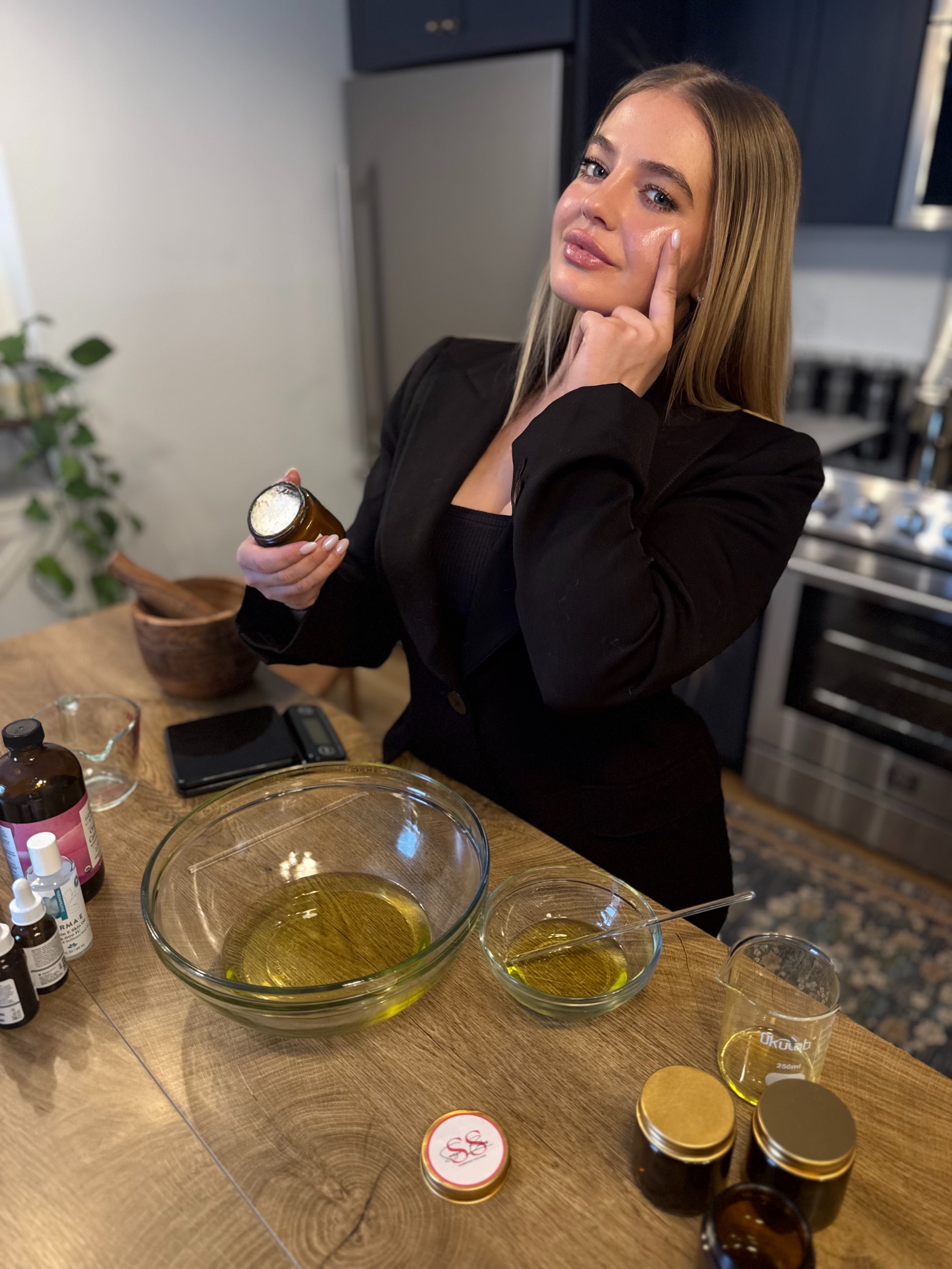 Founder Sabrina Hartz in a kitchen holding a bottle of the Cleopatra beef tallow balm, with various bottles and a bowl on the counter.